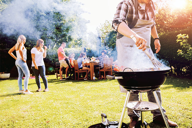 A person grilling food on a barbecue in a sunny backyard while a group of people socialize and sit at a table in the background. Smoke rises from the grill, and trees and grass surround the scene.